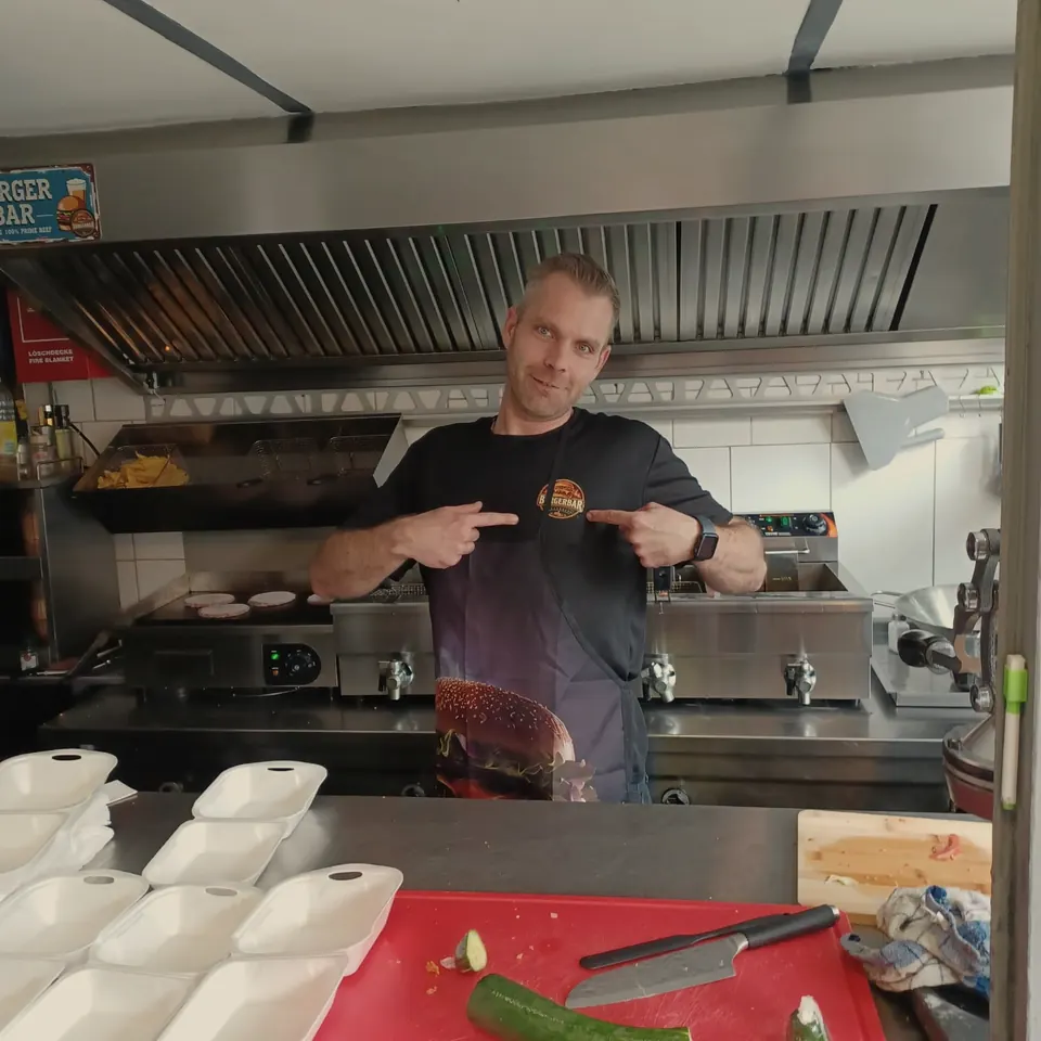 Mike in seiner Küche, zeigt auf das Burgerbar-Logo auf seinem Shirt